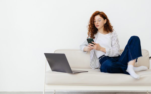 Young beautiful red-haired woman sitting on couch in living room and shopping online on mobile phone with laptop.